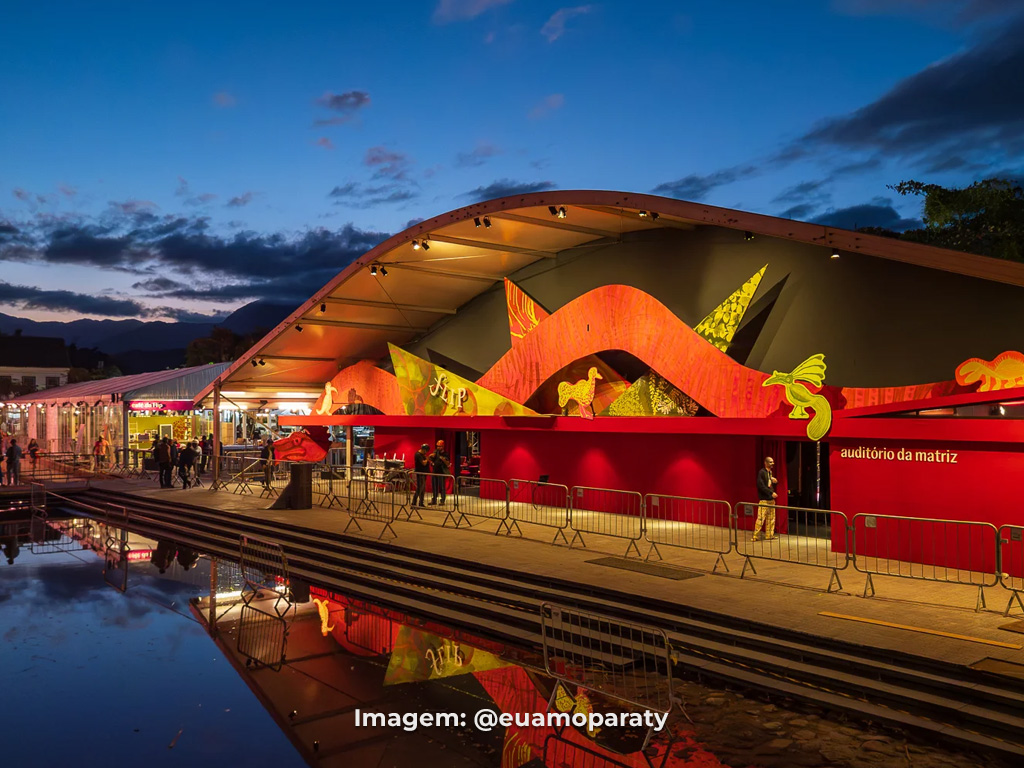 Auditório da Matriz iluminado durante a Flip 2026 em Paraty, com estrutura refletida na água ao entardecer