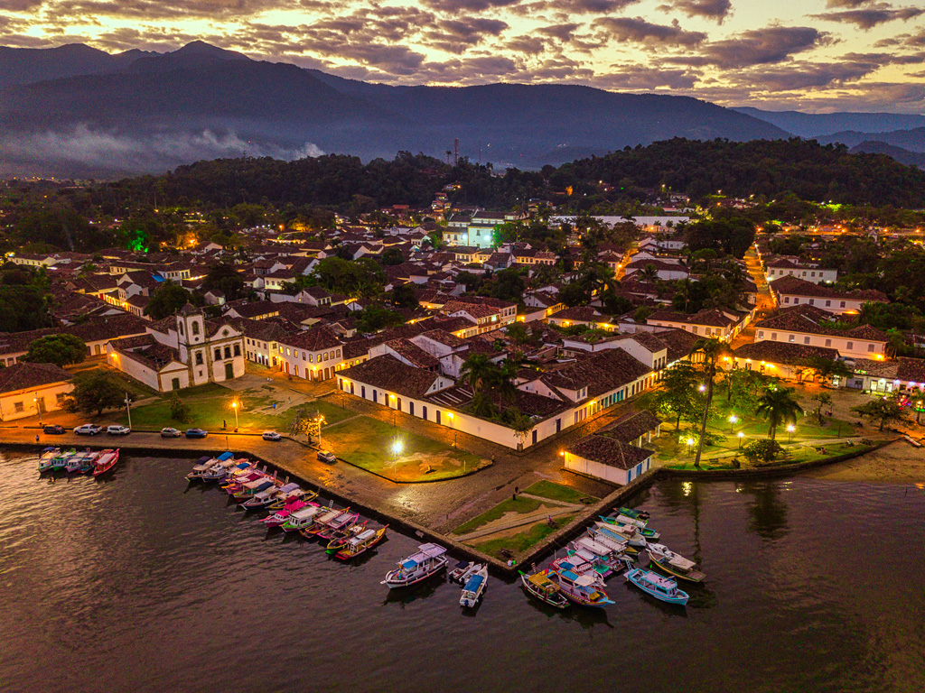 Vista aérea do Centro Histórico de Paraty ao entardecer, com barcos no cais e montanhas ao fundo